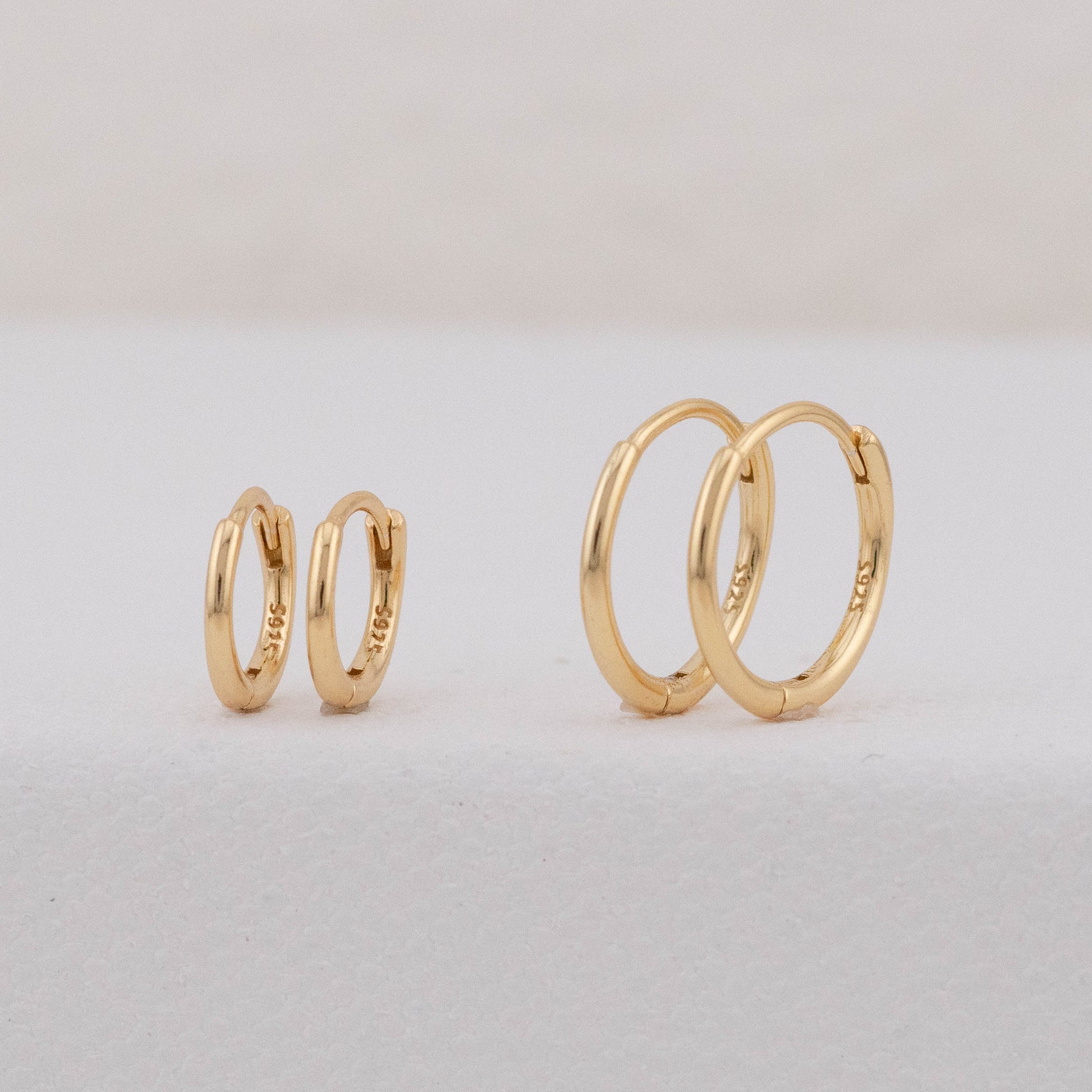 Two pairs of stackable gold huggie hoops with hinged closures in different sizes, displayed upright.